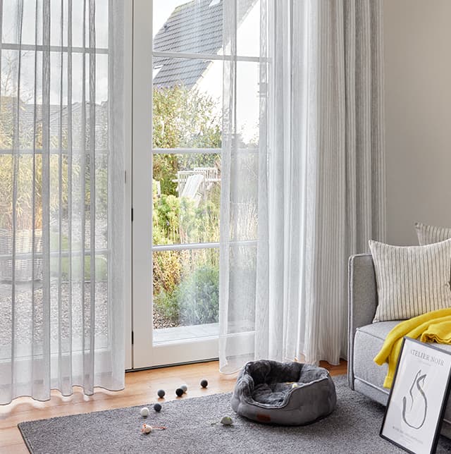 Weiße transparente Gardine am Terrassenfenster und ein Katzenkorb und Katzenspielzeug auf dem Teppich.