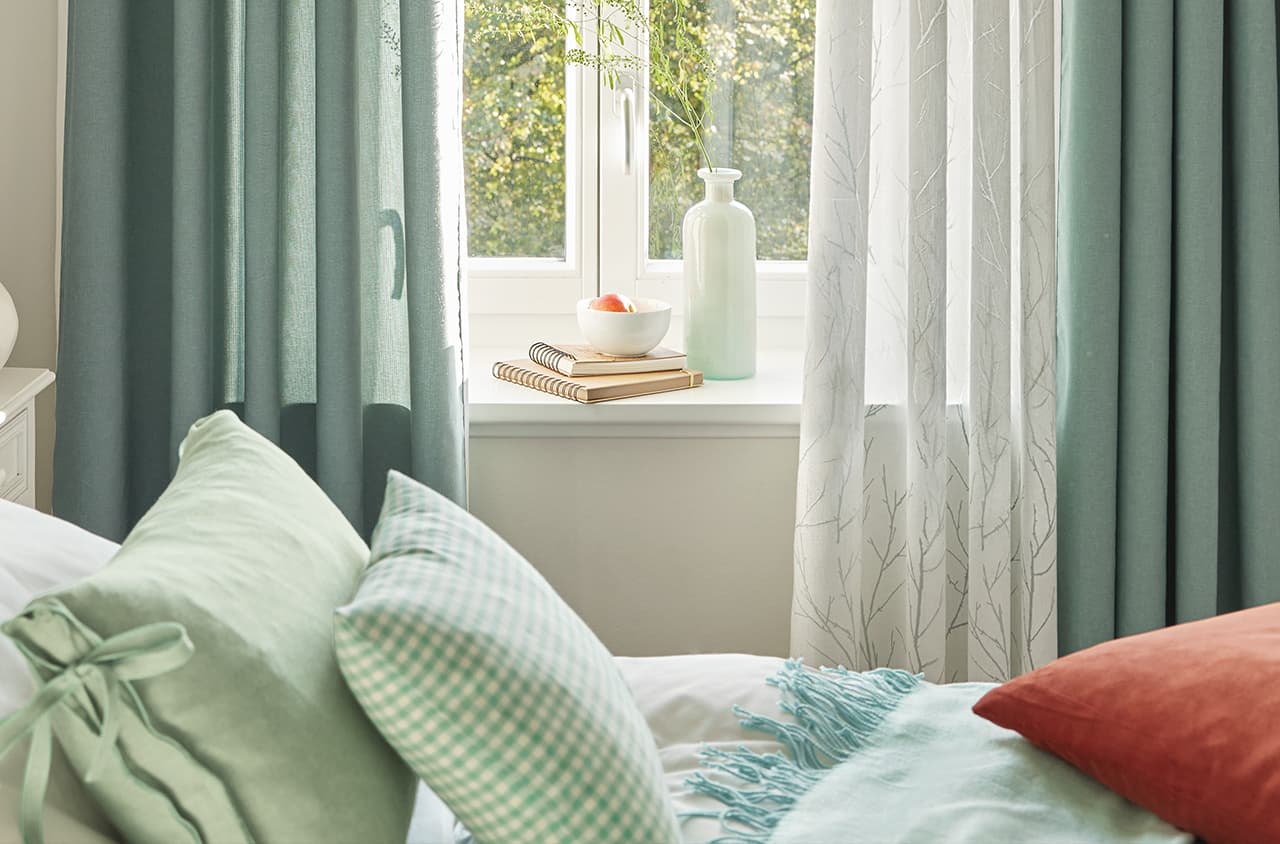 Ein Bett mit zwei grünen und einem roten Kissen vor einem Fenster mit grünen und weißen Gardinen.