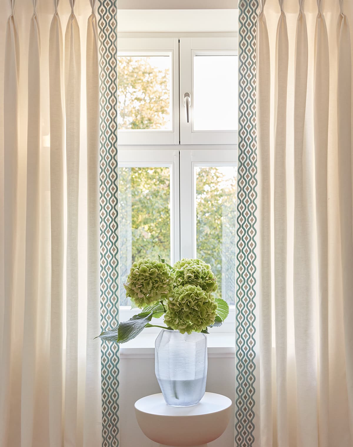 Eine Nahaufnahme von einem beigen Vorhang vor einem Fenster. Im Fokus steht eine Vase mit drei Blüten.