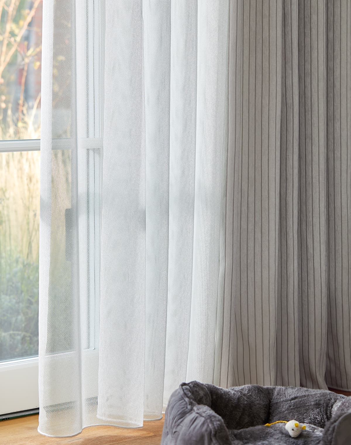 Weiße transparente Gardine am Terrassenfenster und ein Katzenkorb und Katzenspielzeug auf dem Teppich.