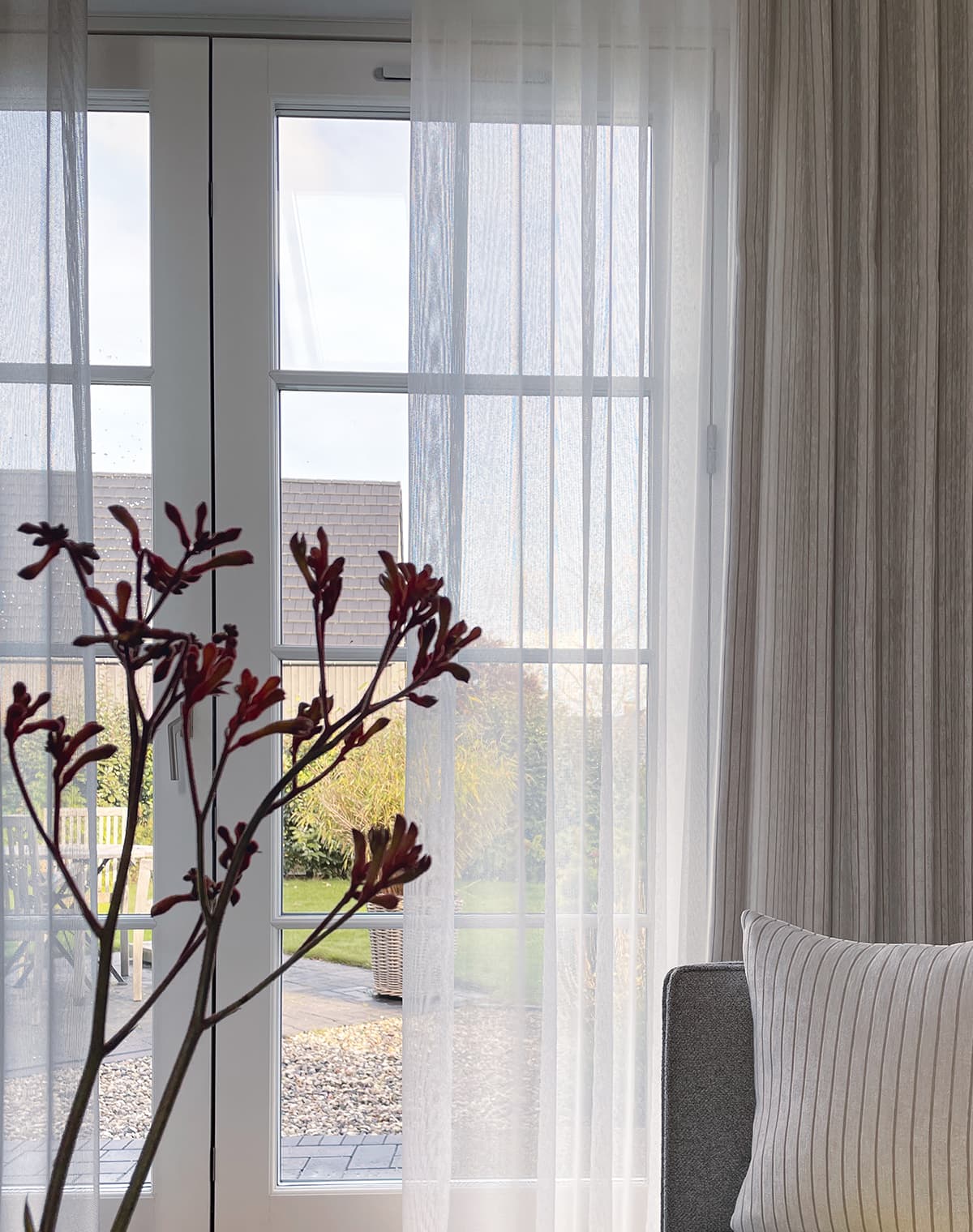 Eine weiße transparente Gardine am Fenster, ein weiß-beige gestreiftes Kissen und eine Blume.