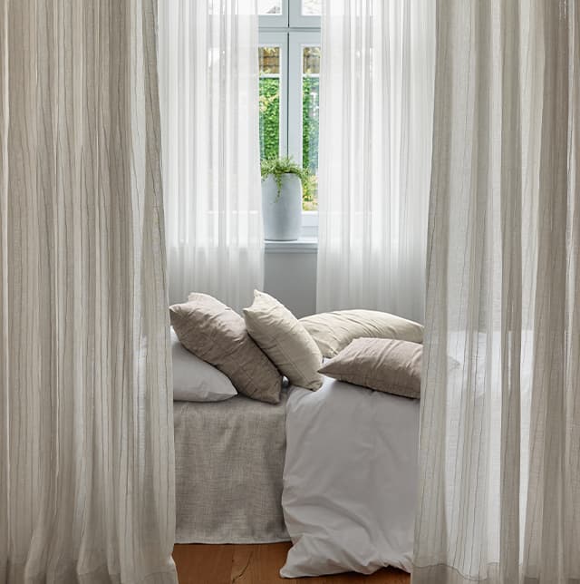 Eine Nahaufnahme von einem Bett auf dem weiß, beige Kissen liegen. Links und rechts von dem Bett sind beige Vorhänge.